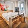 Modernes Hotelzimmer mit Doppelbett, abstraktem Kunstwerk an der Wand und einem Fernseher auf einem Regal.