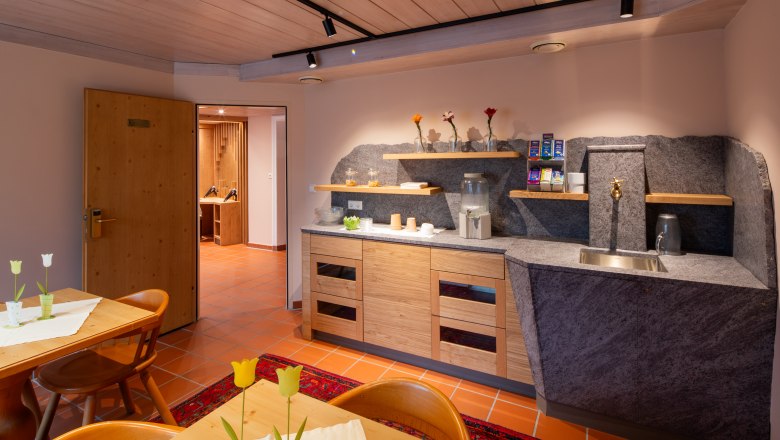 Ein gem&uuml;tlicher Aufenthaltsraum in einem Hotel-Spa mit Holzm&ouml;beln, einer kleinen K&uuml;chenzeile und dekorativen Blumen.