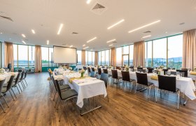 Ein gro&szlig;er, heller Saal mit gedeckten Tischen und St&uuml;hlen, gro&szlig;en Fenstern und einem Projektor.