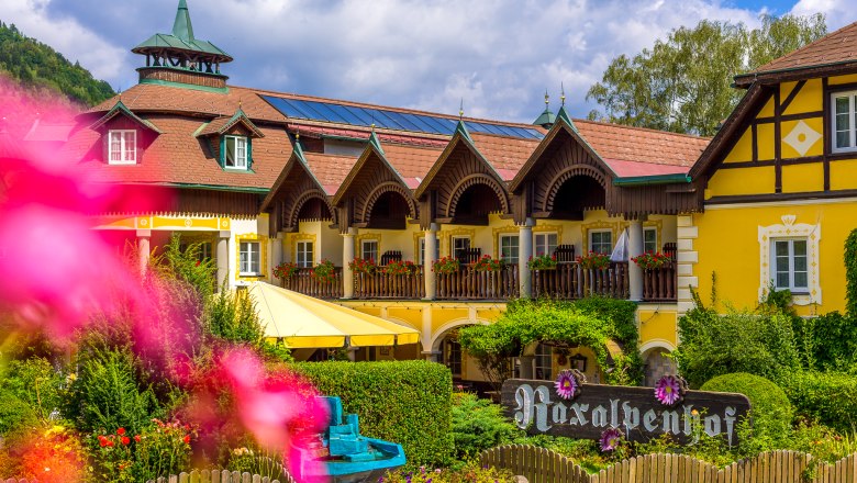 Ein gelbes, traditionelles Hotel mit Holzbalkonen und Blumen, umgeben von gr&uuml;nen B&auml;umen und einem blauen Himmel.