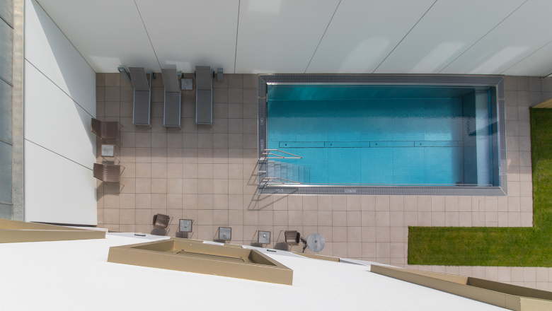 Draufsicht auf einen rechteckigen Pool mit Liegestühlen und Tischen auf einer gefliesten Terrasse.