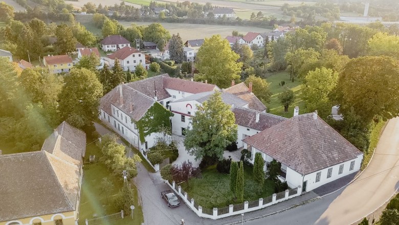 Luftaufnahme eines historischen Gebäudekomplexes mit Innenhof, umgeben von Bäumen und weiteren Häusern in einer ländlichen Gegend.
