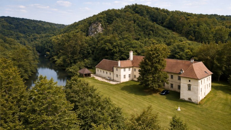 Luftaufnahme eines gro&szlig;en, historischen Geb&auml;udes mit rotem Dach, umgeben von Wald und einem Fluss.