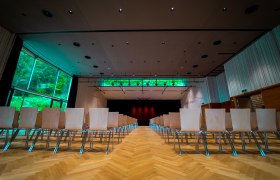 Innenansicht eines modernen Veranstaltungsraums mit leeren Stuhlreihen und einer B&uuml;hne im Hintergrund.