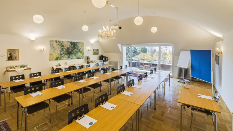 Ein Konferenzraum mit Tischen und St&uuml;hlen, einem Kronleuchter und einem gro&szlig;en Fenster mit Blick nach drau&szlig;en.