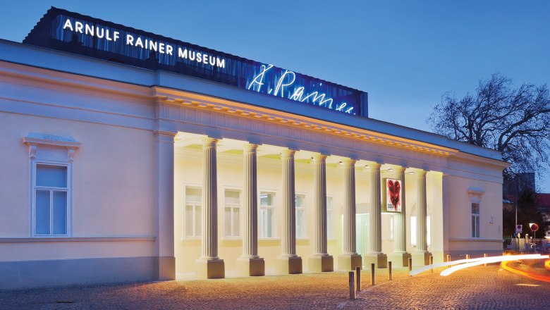 Au&szlig;enansicht des Arnulf Rainer Museums bei D&auml;mmerung mit beleuchteten S&auml;ulen und Leuchtschrift.