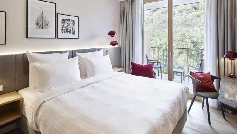 Modernes Hotelzimmer mit gro&szlig;em Bett, Schreibtisch und Balkon mit Aussicht auf gr&uuml;ne Landschaft.