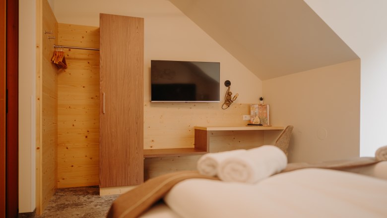 Ein gem&uuml;tliches Hotelzimmer fotografiert vom Bett aus. Gegen&uuml;ber vom Bett h&auml;ngt ein Flachbildfernsehener an der Wand. Daneben ist eine Garderobe aus hellem Holz, die Wand ist auch zum Teil mit Holz verkleidet. Dort befindet sich auch eine Ablagefl&auml;che und ein Schreibtisch mit Sessel und Lampe. Das Zimmer hat eine Dachschr&auml;ge.