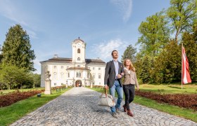 Ein Paar spaziert auf einem gepflasterten Weg vor dem Schlosshotel Rosenau, umgeben von grünen Bäumen und einer österreichischen Flagge.