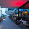 Terrasse eines Restaurants mit Tischen und St&uuml;hlen, beleuchtet von roten Lichtern, mit Blick auf Geb&auml;ude im Hintergrund.