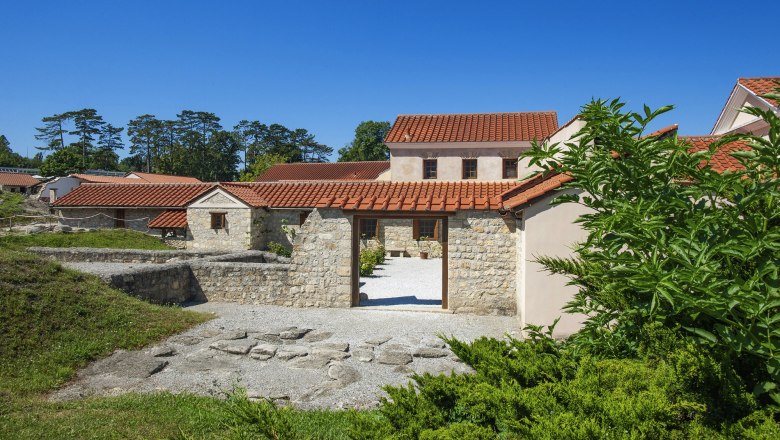 Rekonstruierte römische Gebäude in Carnuntum mit roten Ziegeldächern und Steinmauern, umgeben von grüner Vegetation.
