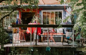 Bunte Terrasse vor der Location mit roten Stühlen und Tisch, umgeben von Pflanzen und Dekorationen.