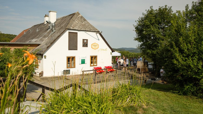 Wei&szlig;es Landhaus mit Schindeldach, umgeben von gr&uuml;ner Landschaft und Menschen im Hintergrund.
