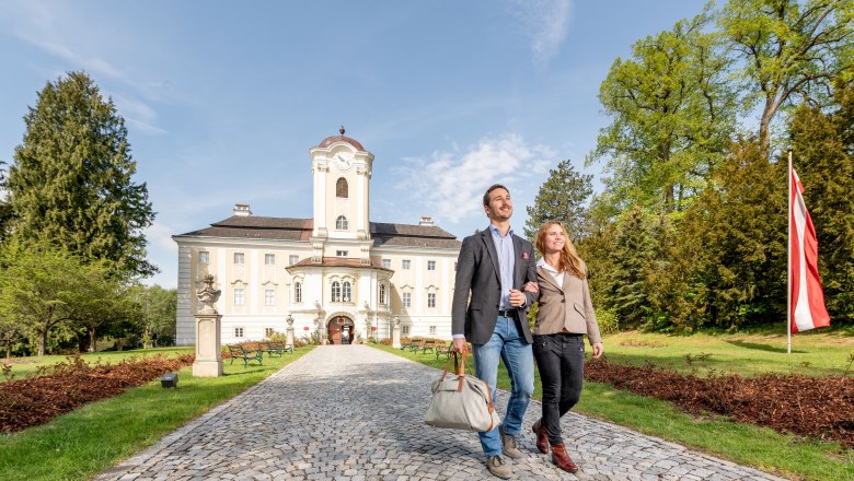 Ein Paar spaziert auf einem gepflasterten Weg vor dem Schlosshotel Rosenau, umgeben von gr&uuml;nen B&auml;umen und einer &ouml;sterreichischen Flagge.