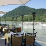 Terrasse mit gedeckten Tischen, Blick auf Fluss und vorbeifahrendes Schiff, umgeben von gr&uuml;nen H&uuml;geln.