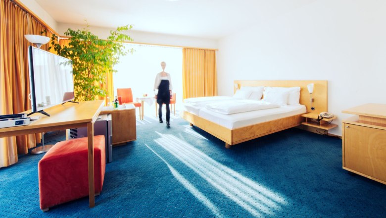 Helles Hotelzimmer mit Doppelbett, Schreibtisch, Fernseher und großer Pflanze. Eine Person geht durch den Raum.