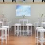 Seminarraum mit wei&szlig;en Stehtischen und Hockern, Blumenvasen und einer Leinwand mit der Aufschrift 'Welcome'.