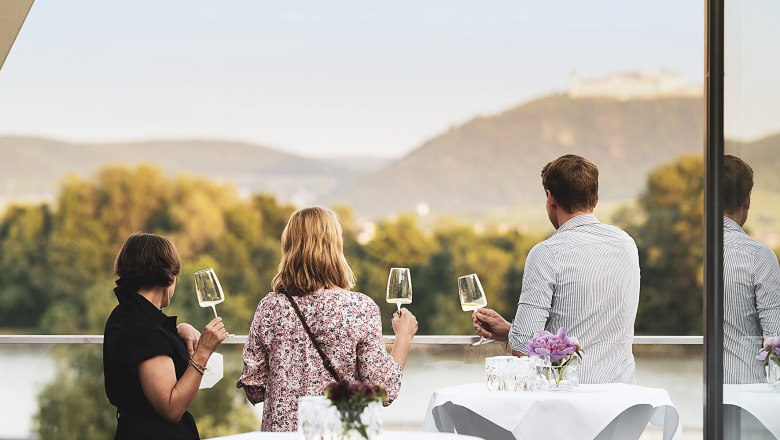 Drei Personen auf einer Terrasse mit Weingl&auml;sern, Blick auf H&uuml;gel und Fluss.
