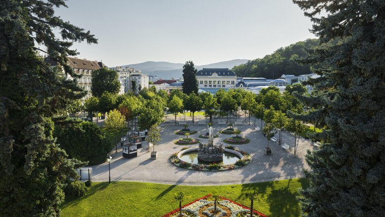 Ein gepflegter Park mit Blumenbeeten, B&auml;umen und einem Brunnen, umgeben von Geb&auml;uden und H&uuml;geln im Hintergrund.