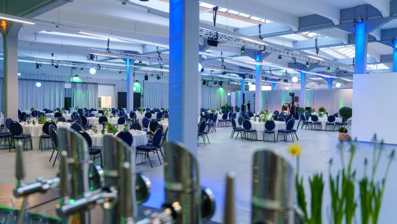 Großer Veranstaltungsraum mit runden, gedeckten Tischen und blauer Beleuchtung.