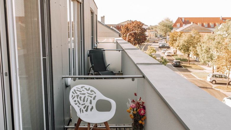 Balkon mit weißem Stuhl und Blumenstrauß, Blick auf Straße und Gebäude.