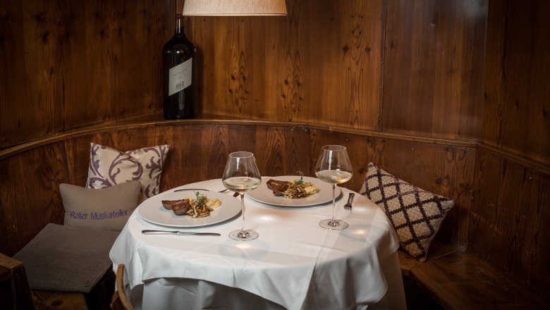 Gem&uuml;tliche Gaststube mit gedecktem Tisch, zwei Tellern mit Speisen und Weingl&auml;sern, Holzwand und Kissen.