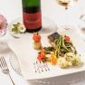 Elegantes Gericht mit Fisch, Spargel und Kartoffeln auf wei&szlig;em Teller, dekoriert mit Blumen. Im Hintergrund eine Flasche Wein und ein Glas.