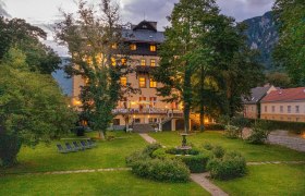 Beleuchtetes Gebäude in einem Garten bei Dämmerung, umgeben von Bäumen und Bergen.