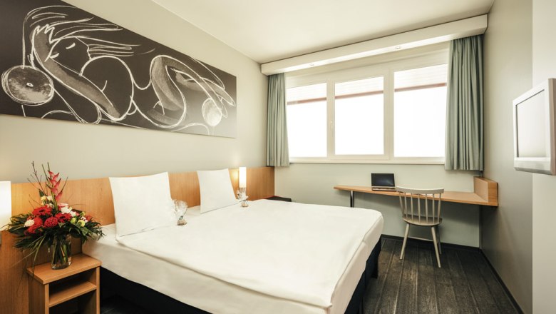 Modernes Doppelzimmer mit gro&szlig;em Bett, Kunstwerk an der Wand, Schreibtisch und Blumenarrangement.