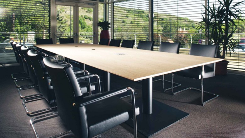 Moderner Seminarraum mit gro&szlig;em Holztisch und schwarzen St&uuml;hlen, umgeben von Glasw&auml;nden mit Blick auf gr&uuml;ne Landschaft.