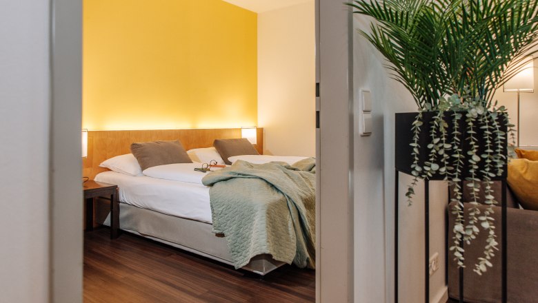 Hotelzimmer mit gelber Wand, Doppelbett und Pflanze im Vordergrund.