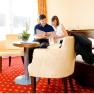 Ein Paar sitzt in einem Hotelzimmer auf einem Bett und schaut in ein Buch. Der Raum hat rote Teppiche und gelbe Vorh&auml;nge.