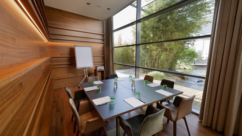 Moderner Seminarraum mit Holzw&auml;nden, gro&szlig;em Tisch, St&uuml;hlen und Flipchart. Gro&szlig;e Fenster mit Blick auf Garten.