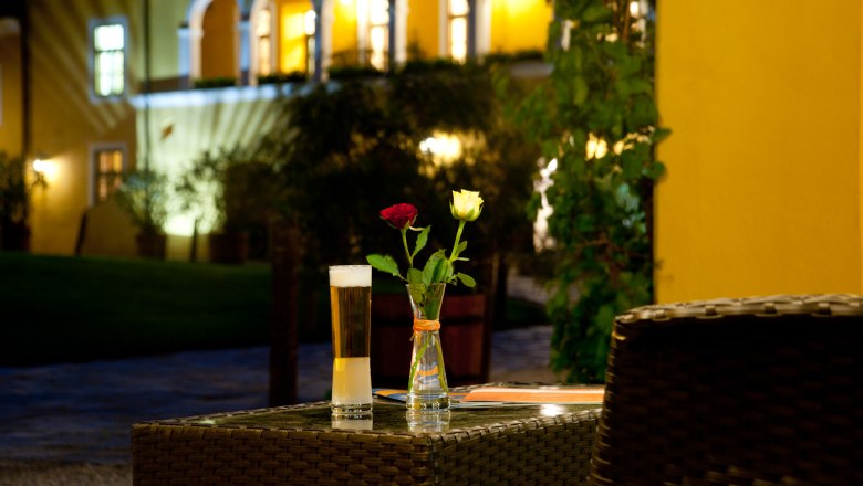 Ein Tisch mit einem Glas Bier und zwei Rosen in einer Vase, im Hintergrund ein beleuchtetes Geb&auml;ude mit B&ouml;gen.