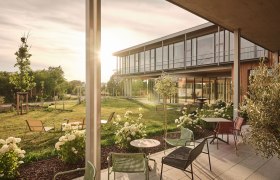 Moderne Terrasse mit Gartenm&ouml;beln und Blumen vor einem Geb&auml;ude bei Sonnenuntergang.
