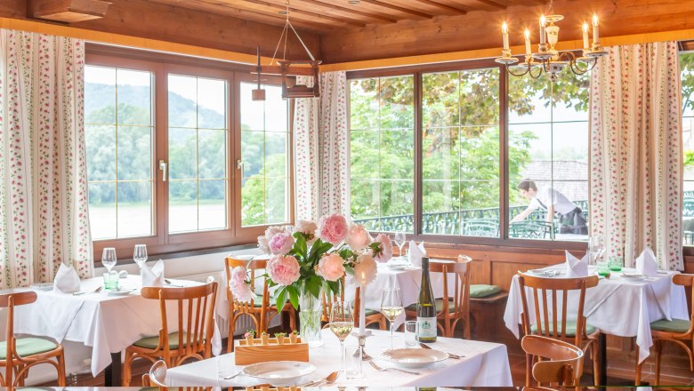 Elegantes Restaurant mit gedeckten Tischen, Blumen und Blick auf die Natur durch gro&szlig;e Fenster.
