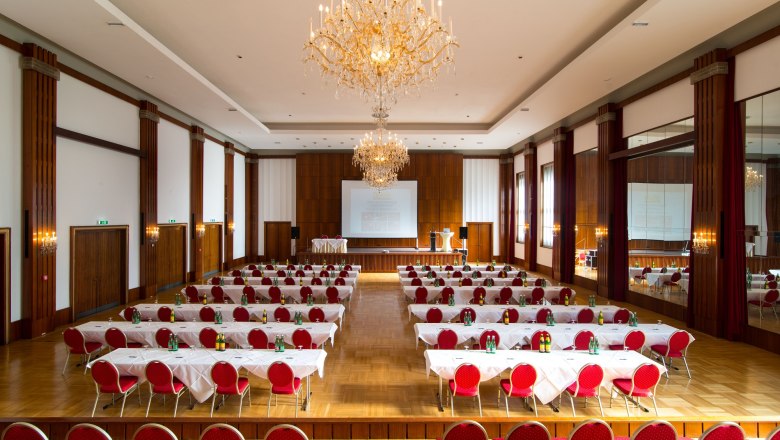 Ein großer, eleganter Konferenzsaal mit roten Stühlen, weißen Tischdecken und Kronleuchtern.