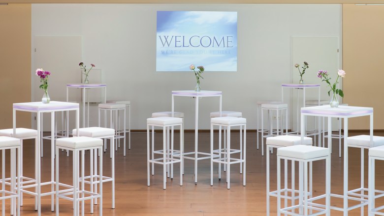 Seminarraum mit wei&szlig;en Stehtischen und Hockern, Blumenvasen und einer Leinwand mit der Aufschrift 'Welcome'.