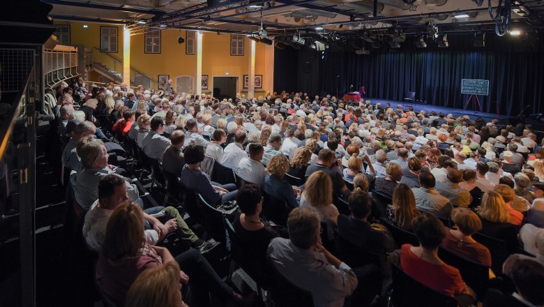 Ein gef&uuml;llter Theatersaal mit Publikum, das auf eine B&uuml;hne blickt.