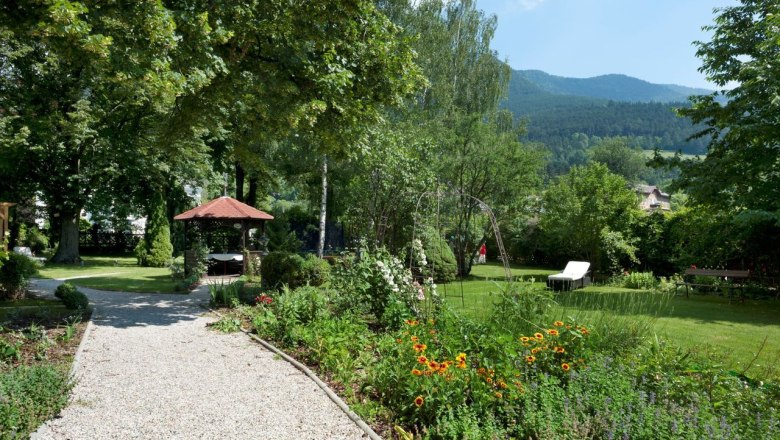 Ein gepflegter Garten mit Kiesweg, Blumenbeeten, einem Pavillon und einer Liege. Im Hintergrund sind B&auml;ume und Berge zu sehen.