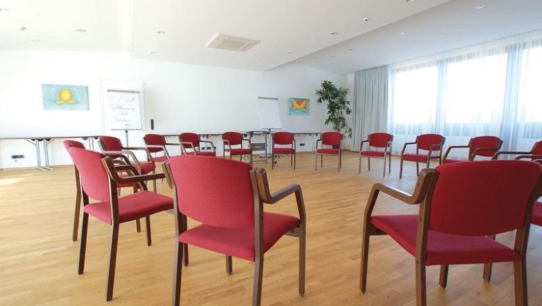 Ein heller Seminarraum mit roten St&uuml;hlen im Kreis, Holzboden und gro&szlig;en Fenstern.