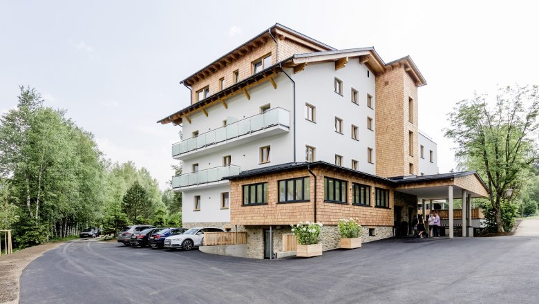 Ein modernes Hotelgebäude mit Holzverkleidung und Balkonen, umgeben von Bäumen und einem Parkplatz mit Autos.