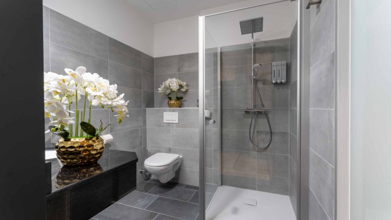 Modernes Badezimmer mit grauen Fliesen, Dusche, Toilette und Orchideen in goldenen Vasen.