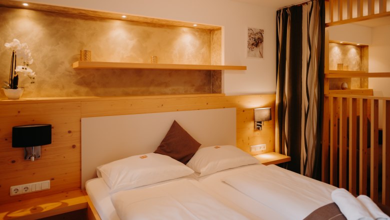 Gem&uuml;tliches Hotelzimmer mit Doppelbett, heller Holzverkleidung, Raumtrenner aus Holz und mit einem Vorhang. Neben dem Bett sind Steckdosen und Lichtschalter. &Uuml;ber dem Bett eine Nische in der Wand, die beleuchtet und in einem erdigen Goldton gestrichen ist.