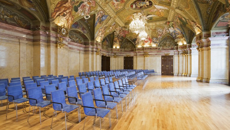 Innenansicht des Landtagssaals im Palais Nieder&ouml;sterreich mit blauen St&uuml;hlen und kunstvoll bemalter Decke.