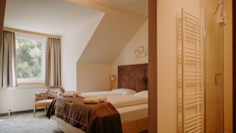 Gem&uuml;tliches Hotelzimmer mit Doppelbett aus hellem Holz, Fenster mit Vorh&auml;ngen und einem Sessel. Im Vordergrund sieht man ins Badezimmer. Das Zimmer hat fensterseitig eine Dachschr&auml;ge.