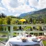 Blick von einem Balkon mit Tisch, Blume, Getr&auml;nken und Buch, im Hintergrund Berge und gr&uuml;ne Landschaft.