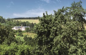 Ein Hotelgeb&auml;ude inmitten einer gr&uuml;nen Landschaft mit B&auml;umen und H&uuml;geln im Hintergrund.