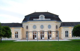 Historisches Gebäude mit symmetrischer Fassade und großen Fenstern, umgeben von Grünflächen.