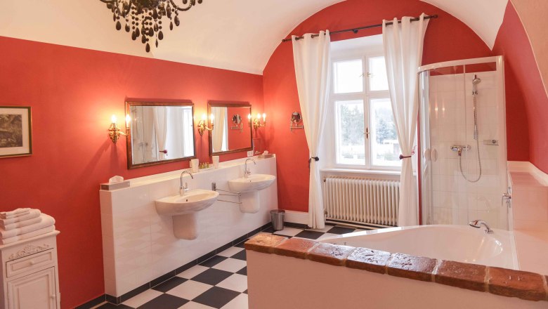 Elegantes Badezimmer mit roten W&auml;nden, zwei Waschbecken, Badewanne, Dusche und Kronleuchter.